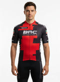 Male cyclist riding a road bike down a mountain highway wearing a black and red short sleeve BMC Switzerland cycling jersey and matching bib shorts.