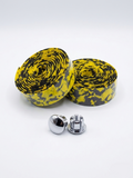 Two rolls of premium yellow and black splatter patterned bicycle handlebar tape displayed together with two metallic chrome handlebar end plugs on a clean white background